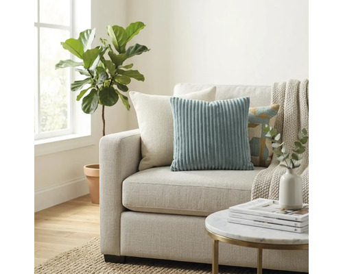 Ljust vardagsrum med tygsoffa, prydnadskuddar i manchester, en pläd, ett runt sidobord och en stor inomhusväxt intill fönstret.
