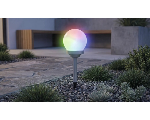 Solcellsdriven globformad trädgårdsbelysning med flerfärgad RGB-belysning, på ett markspett i en grusbädd med växter.