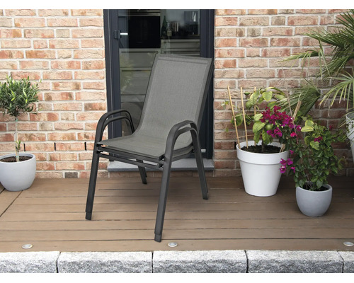 Stapelbar trädgårdsstol med aluminiumram och textilsits på en terrass med trädäck och växter framför en tegelvägg.