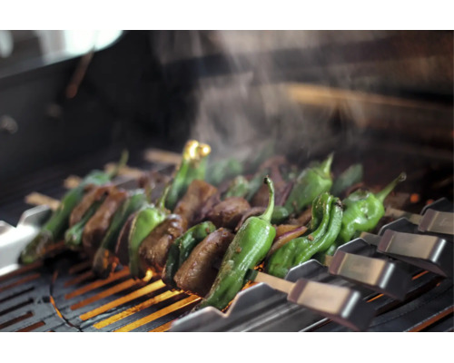 Kött och grön paprika på metallspett över ett varmt grillgaller med stigande ånga.
