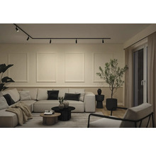 Modernt vardagsrum med skensystem i taket, en stor modulsoffa med kuddar och vägglister. En hög krukväxt och små soffbord kompletterar den ljusa och minimalistiska inredningen.