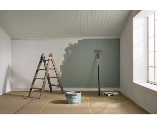 Renovering av ett rum med en vägg delvis målad i grågrönt. Trästege, färghink, roller och täckpapper på golvet syns.