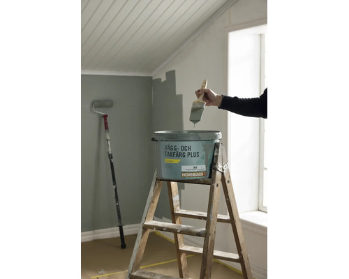 En hink Hornbach vägg- och takfärg på en trästege med en hand som håller en pensel ovanför.
