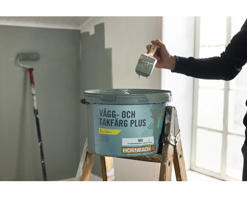 En person håller en målarpensel över en öppen hink Hornbach vägg- och takfärg som står på en trappstege av trä i ett ljust rum.