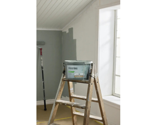 En hink Hornbach väggfärg på en trappstege i trä i ett ljust rum med en delvis målad vägg.
