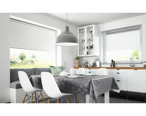 Ljust kök och matplats med mönstrad duk, vita stolar, grå taklampa och fönster med persienner.