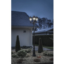 Trearmad utomhuslykta i klassisk design, upplyst i en anlagd trädgård med grus och buskar framför ett bostadshus.