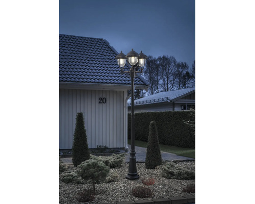 Trearmad utomhuslykta i klassisk design, upplyst i en anlagd trädgård med grus och buskar framför ett bostadshus.