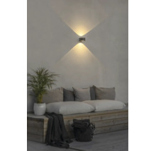Modern LED-vägglampa med ljuseffekt uppåt och nedåt på en vägg ovanför en träbänk med kuddar och en växt.