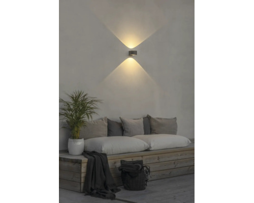 Modern LED-vägglampa med ljuseffekt uppåt och nedåt på en vägg ovanför en träbänk med kuddar och en växt.