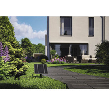 Solcellsdriven trädgårdsspotlight med solpanel och markspot vid en stenlagd gång, med ett modernt hus och en uteplats i bakgrunden.