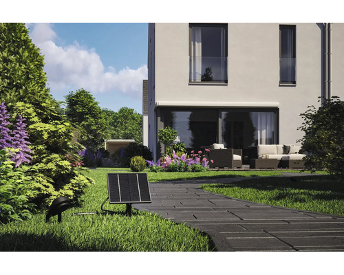 Solcellsdriven trädgårdsspotlight med solpanel och markspot vid en stenlagd gång, med ett modernt hus och en uteplats i bakgrunden.