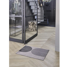 Modern dörrmatta med geometriskt mönster på fiskbensparkett i en ljus hall med glasdörr och trappa.