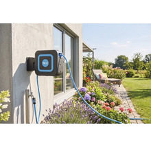 Väggmonterad slangbox med for.q Logo på en husvägg i en trädgård med blombänkar, vattenanslutning, uteplats och gräsmatta.