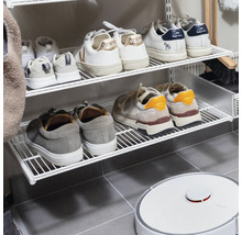 Väggmonterat skoställ i vit tråd med sneakers och en vit robotdammsugare på ett grått klinkergolv.