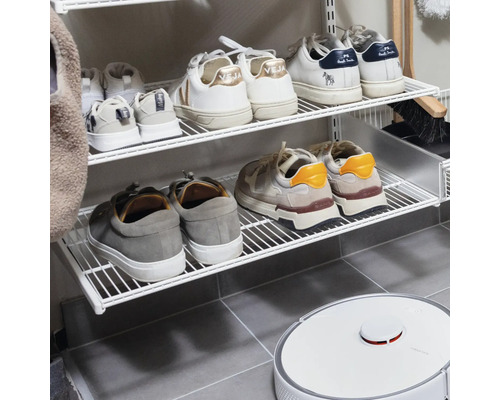 Väggmonterat skoställ i vit tråd med sneakers och en vit robotdammsugare på ett grått klinkergolv.