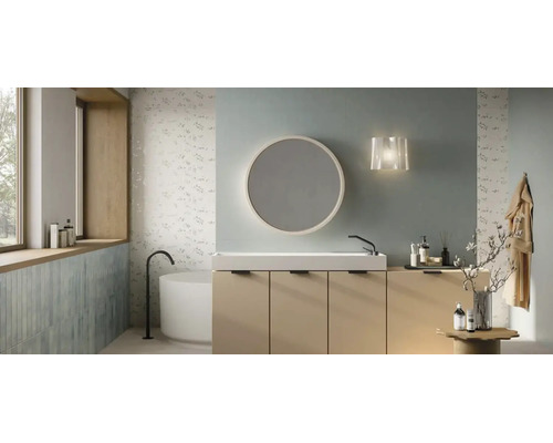 Modernt badrum med beige kommod, vitt tvättställ, rund spegel och fristående badkar framför kakel med blommotiv.