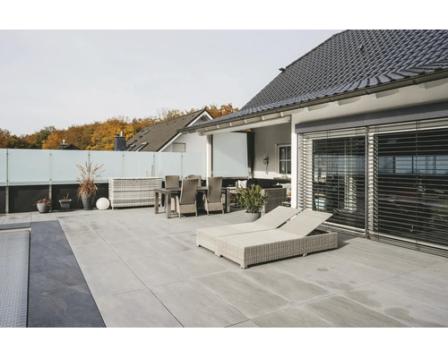 Modern terrass med ljusgrå golvplattor, ljusa rottingträdgårdsmöbler, solsängar och insynsskydd av glas.