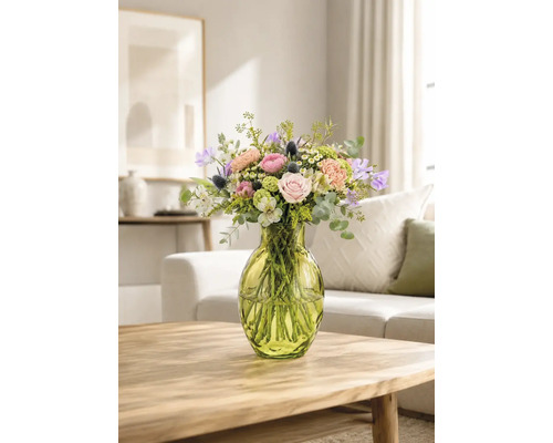 Färgglad blombukett med rosor och eukalyptus i en grön glasvas på ett träbord i ett ljust vardagsrum.