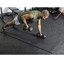 En man utför styrketräning med hantlar på en svart fitnessmatta i ett ljust, organiserat gym med gummigolv.