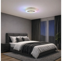 Modern takfläkt med färgad LED-ljusring i ett sovrum med grå klädd säng, mörka gardiner och matta.