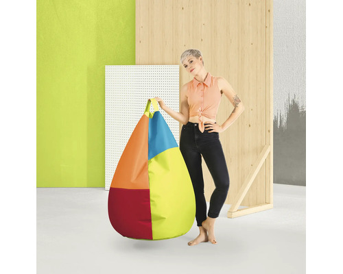 En kvinna står bredvid en stor, droppformad, flerfärgad saccosäck med handtag i en modern studiomiljö med träelement.