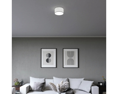 Modern cylindrisk taklampa i ett minimalistiskt vardagsrum med soffa och abstrakta tavlor på en grå vägg.