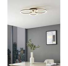 Modern LED-taklampa med fyra överlappande ljusringar i ett ljust rum med grå väggar, bord med vas och stadsutsikt.
