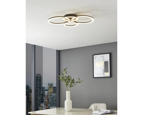 Modern LED-taklampa med fyra överlappande ljusringar i ett ljust rum med grå väggar, bord med vas och stadsutsikt.