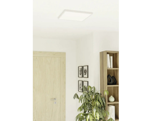 Platt, kvadratisk LED-panel i taket i ett ljust, modernt inrett rum med trädörr, bokhylla och rumsväxt.