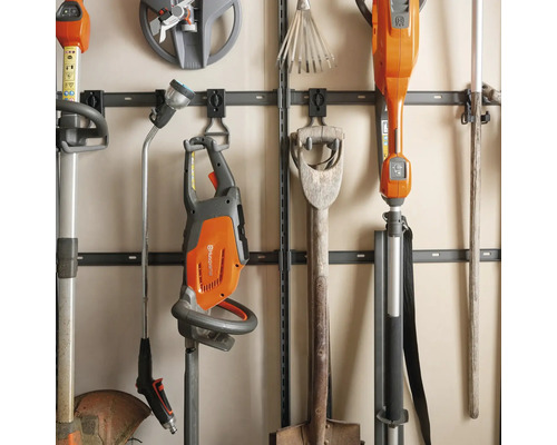 Organiserade trädgårdsredskap på en väggskena med Husqvarna logotyp, inklusive häcksax, röjsåg, spade och sprutlans.