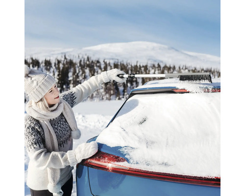 Person tar bort snö från ett biltak med en utdragbar snöborste i ett vinterlandskap.