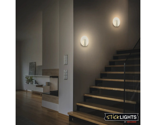 Två runda LED vägglampor ger indirekt ljus i en modern trappa med trätrappsteg och Briloner Sticklights logotyp.