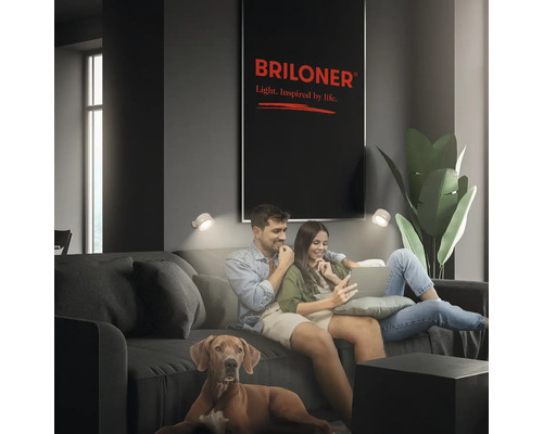 Par sitter med hund i en soffa i ett modernt inrett vardagsrum med vägglampor och en Briloner Leuchten logotyp på väggen.
