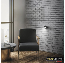 Vägglampa Sticklights från Briloner i ett modernt vardagsrum med tegelvägg och fåtölj för riktad accentbelysning.