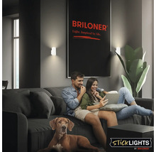 Vardagsrum med par och hund i soffa, upplyst av två vägglampor från Briloner. Briloner logotyp och Sticklights logotyp syns.