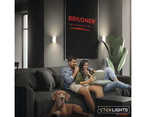 Vardagsrum med par och hund i soffa, upplyst av två vägglampor från Briloner. Briloner logotyp och Sticklights logotyp syns.