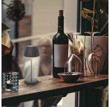 En liten, modern batteridriven LED bordslampa med plisserad skärm på en träbar bredvid en vinflaska och vinglas.
