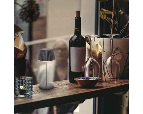 En liten, modern batteridriven LED bordslampa med plisserad skärm på en träbar bredvid en vinflaska och vinglas.