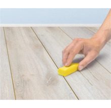 En hand använder ett litet gult block för att rengöra fogen mellan brädorna på ett ljust laminatgolv.