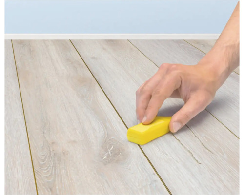 En hand använder ett litet gult block för att rengöra fogen mellan brädorna på ett ljust laminatgolv.