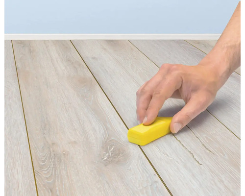 En hand använder en reparationsvaxstift för att laga repor på ett laminatgolv.