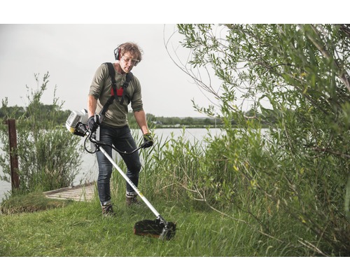 Man använder en grästrimmer med bärrem och skyddsutrustning i en trädgård vid en sjö