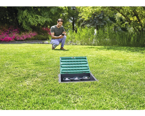 Trädgårdslandskap med en man som använder en smartphone, i förgrunden en strömfördelningsbox inbäddad i gräsmattan med ett öppet lock.