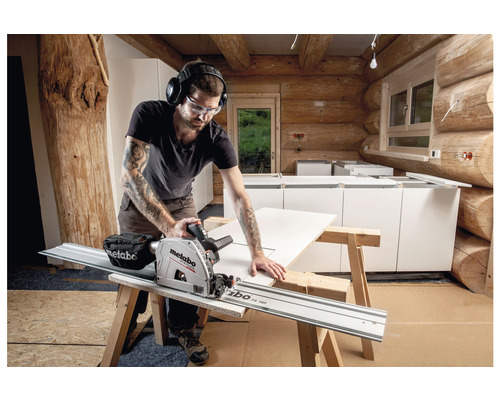 En hantverkare med skyddsglasögon och hörselskydd sågar en skiva med en Metabo sänksåg i ett rum med träväggar.