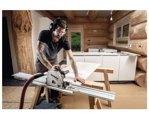 En man skär ett arbetsstycke med en sänksåg och styrskena i ett timmerhus. Han bär skyddsglasögon och hörselkåpor.