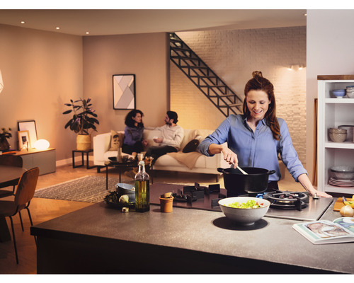 Ljust, öppet kök med en kvinna som lagar mat vid spisen medan två personer sitter i en soffa