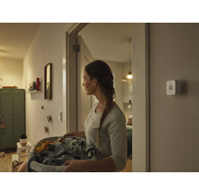 En kvinna bär en tvättkorg genom en hall, på väggen sitter en smart rörelsesensor monterad.