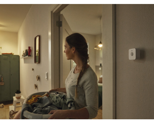 En kvinna bär en tvättkorg genom en hall, på väggen sitter en smart rörelsesensor monterad.