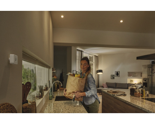 Vit rörelsesensor på en vägg i ett modernt kök med en kvinna som bär en matkasse.